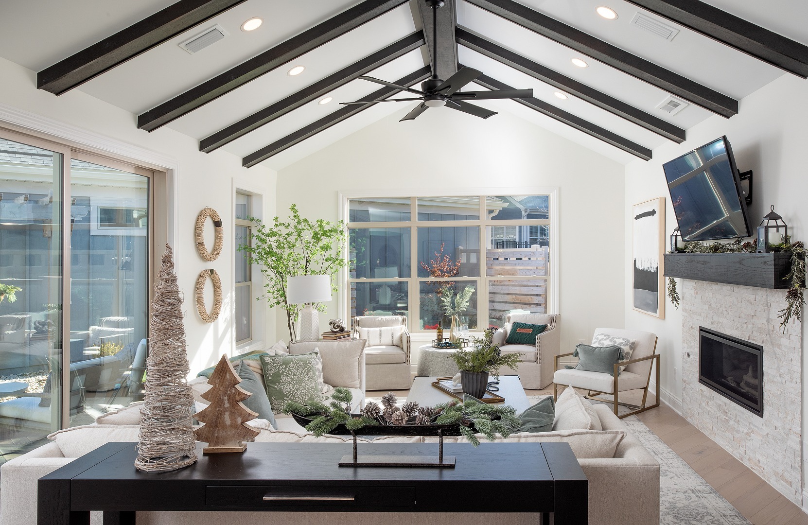 Bright living room with vaulted ceiling, large windows, neutral furniture, holiday decor, and exposed dark wood beams.