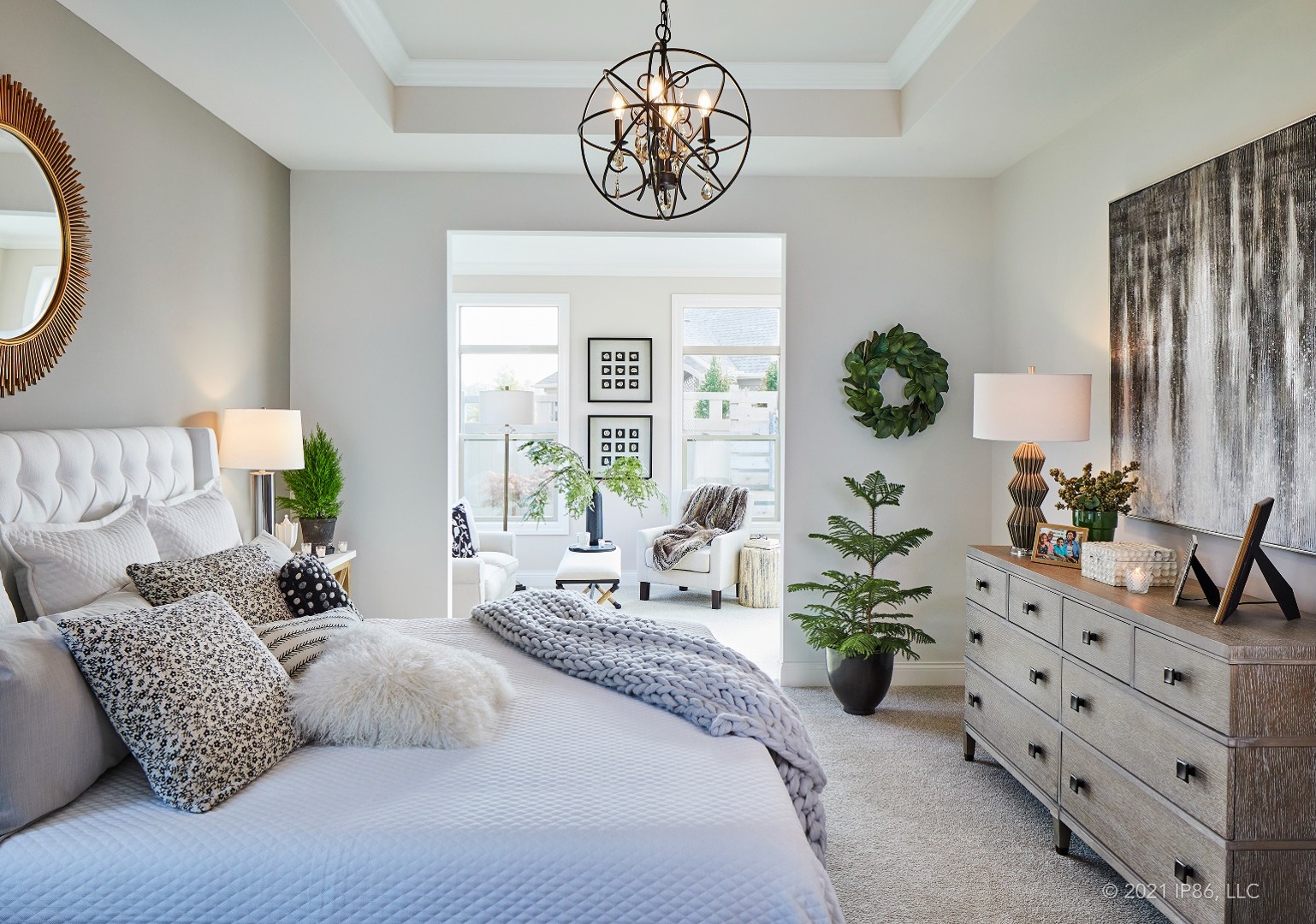 Bright bedroom with neutral tones, cozy bedding, modern chandelier, greenery accents, and a sitting area with large windows.